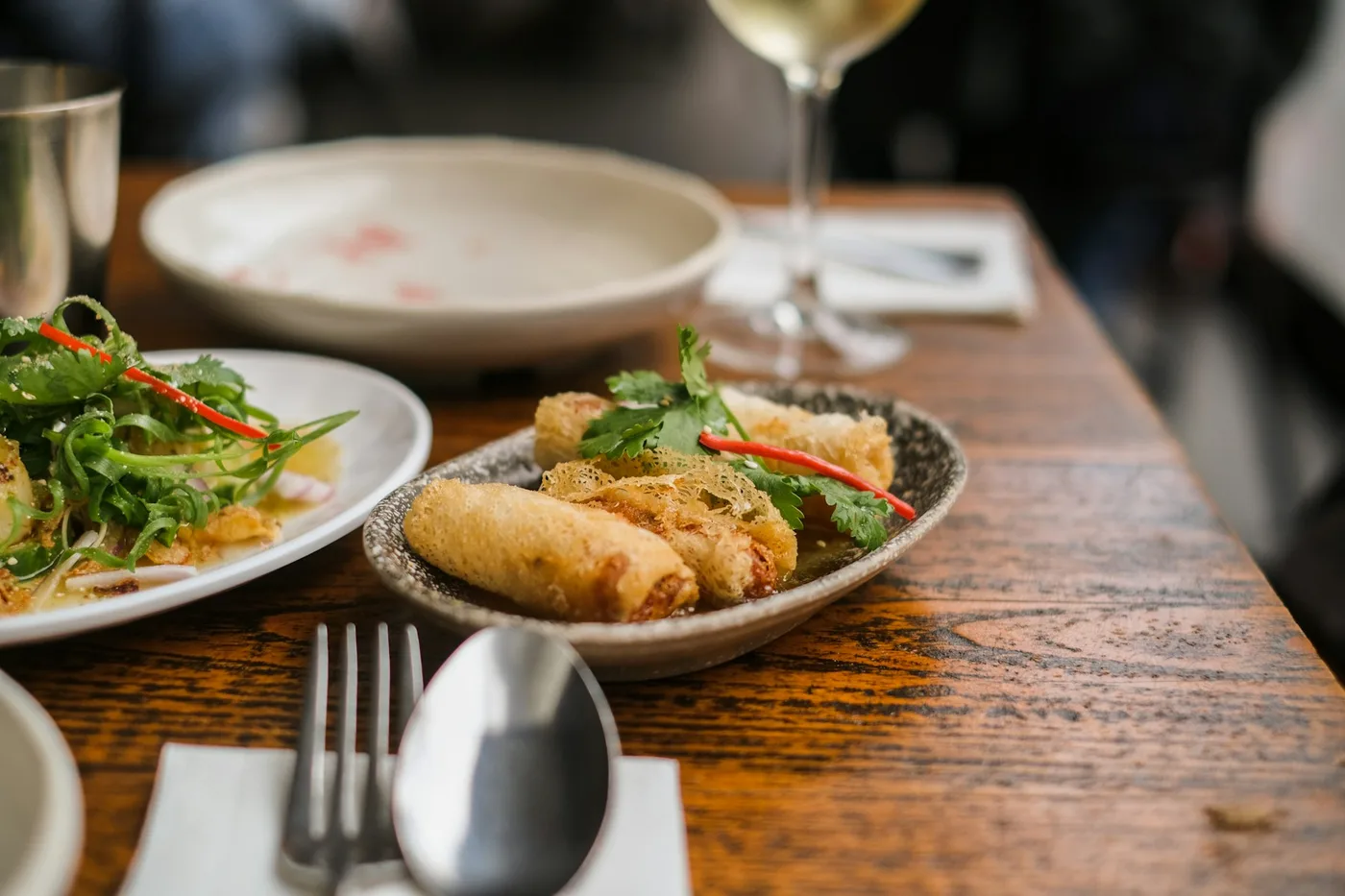 Crispy spring rolls and a herb salad on a restaurant table with a glass of white wine